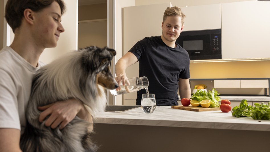 Two boys and a dog are in the kitchen. One of the boys is pouring water into a glass.