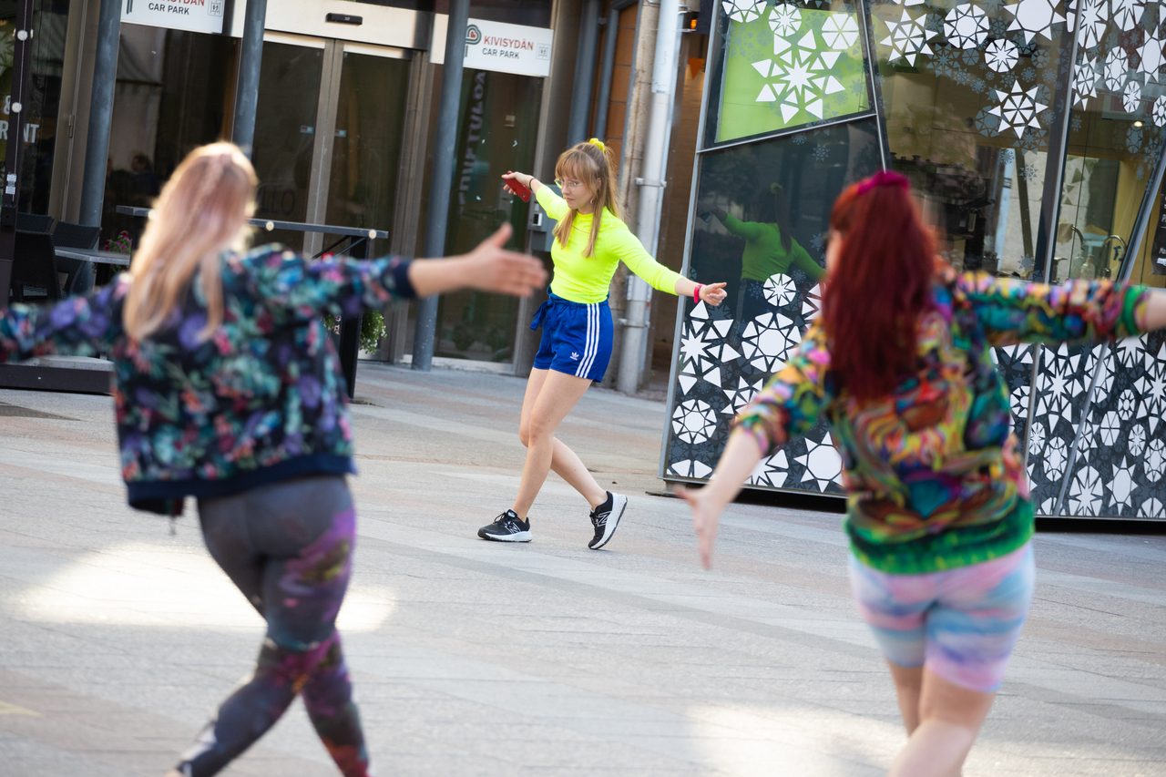 Keltaiseen paitaan pukeutunut henkilö tanssii kädet sivuilla Oulun keskustassa, reunoilla kaksi henkilöä tanssii sellin.