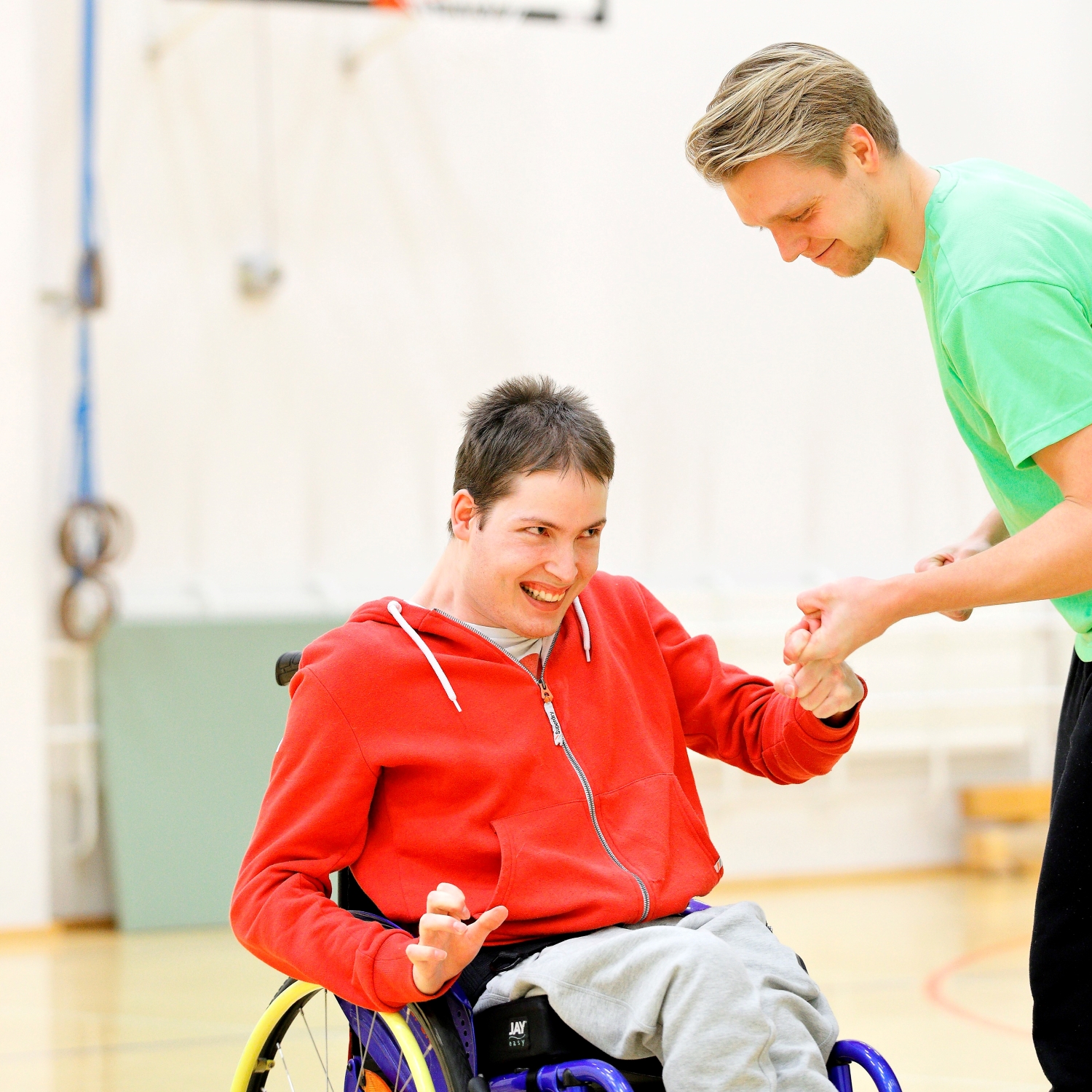 Pyötätuolissa istuva poika pitää kädestä kiinni ohjaajaa liikuntasalissa.