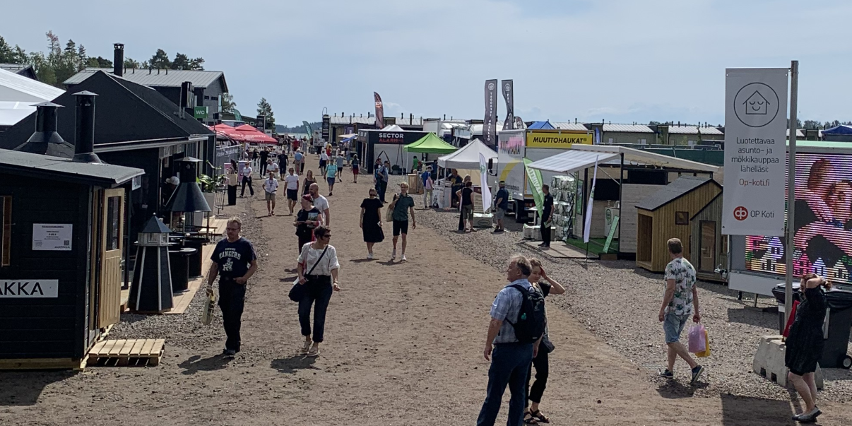 Kuvassa näkyy taivasta ja nurmialuetta sekä esittelykojuja ja ihmisiä soratien varrella.