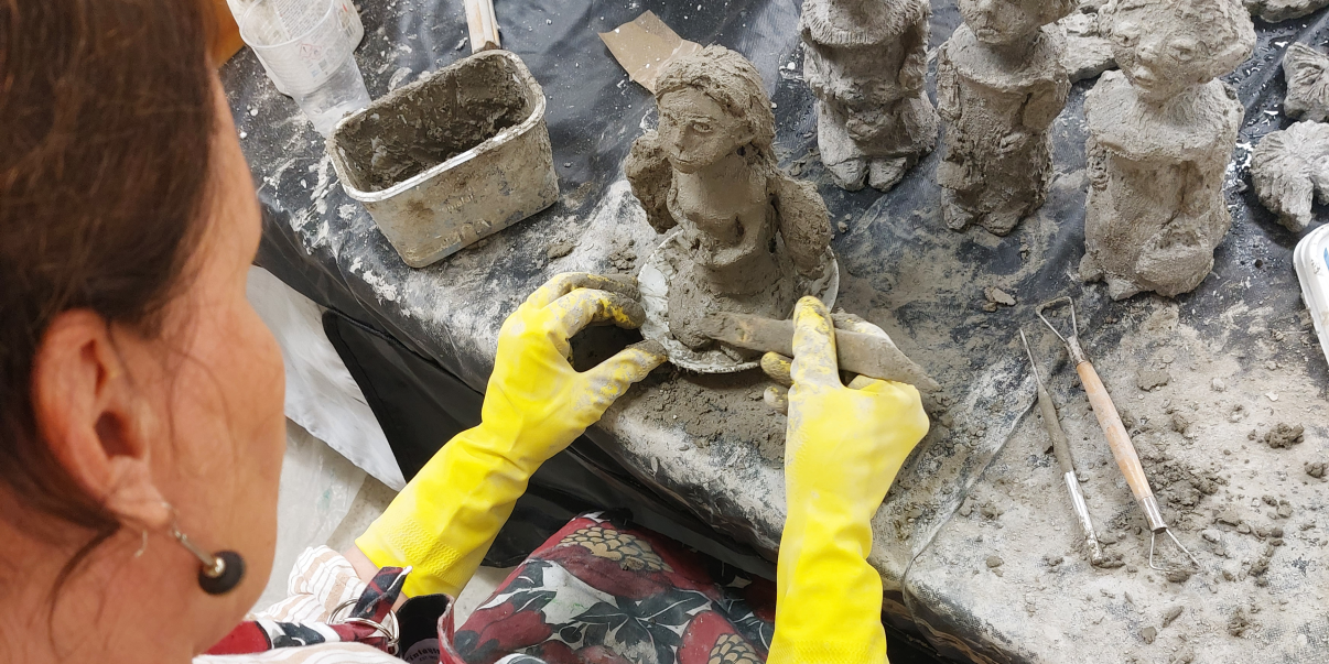 Taiteilija työstää savesta isopäisiä ihmishahmoja keltaiset kumihanskat käsissään.