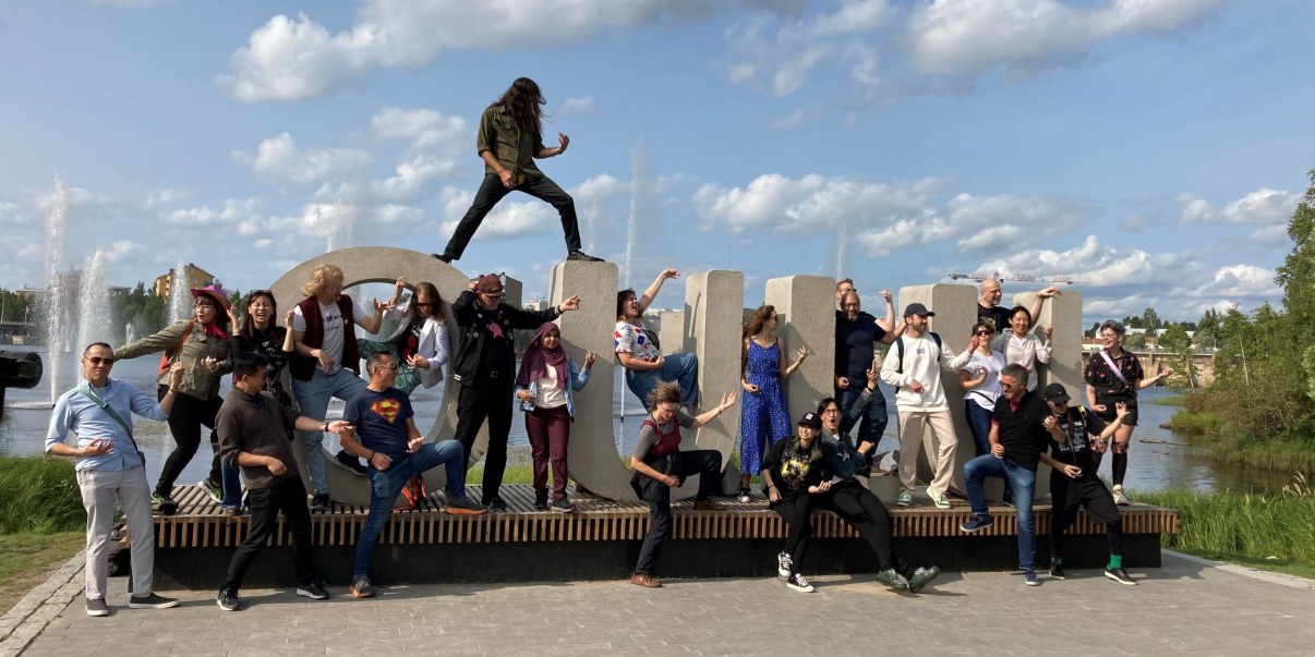 Betonisten OULU -kirjainten läheisyydessä, välissä ja päällä ihmisiä. Soittavat ilmakitaraa, riehakas tunnelma aurinkoisella säällä. Ainolan puiston suihkulähteet taustalla. 