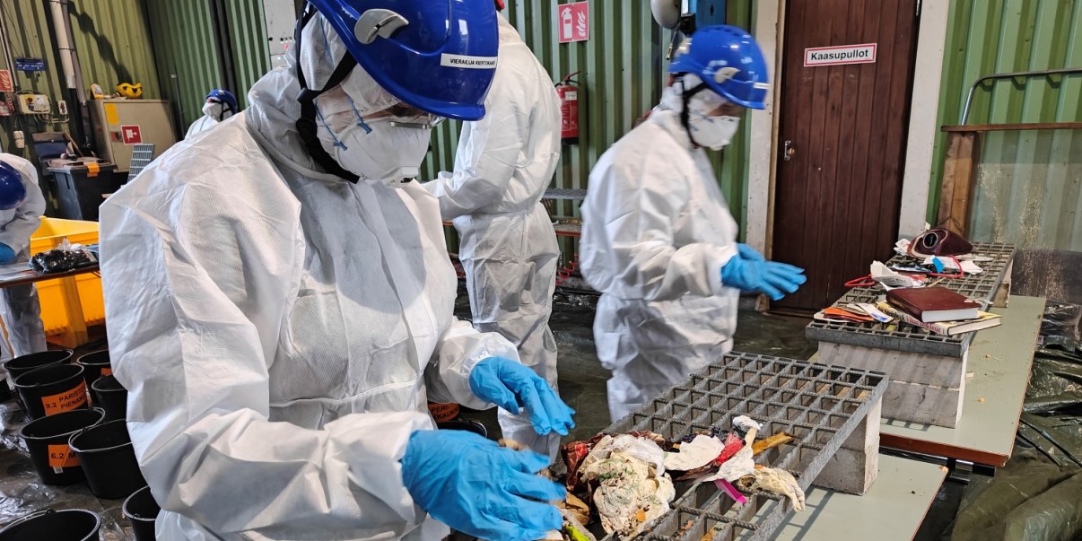 Kolme ihmistä valkoisissa suoja-asuissa ja sinisissä kypärissä. Etualalla olevan ihmisen edessä roskapussin sisältö tutkittavana.