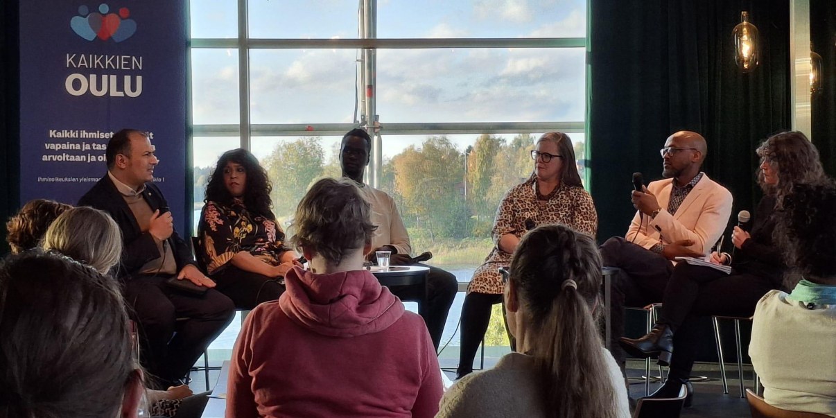 Kuvassa on paljon erilaisia ihmisiä. Lavalla on kuusi ihmistä keskustelemassa.