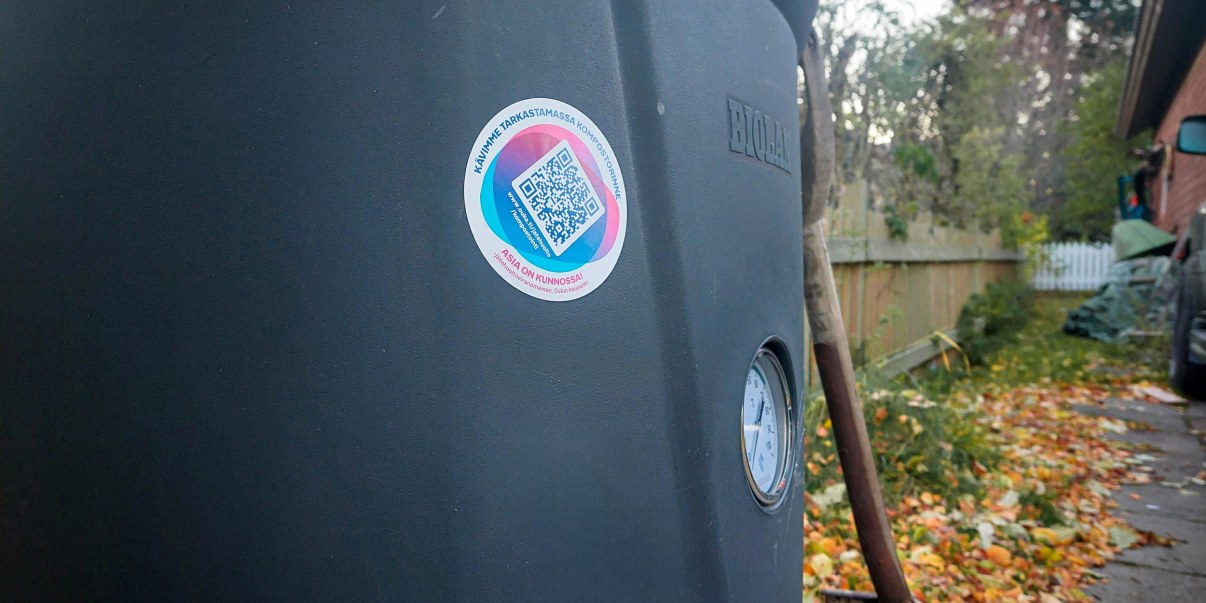 Musta komposti, jonka kyljessä tarra. Lapio nojaa vasten kompostia.