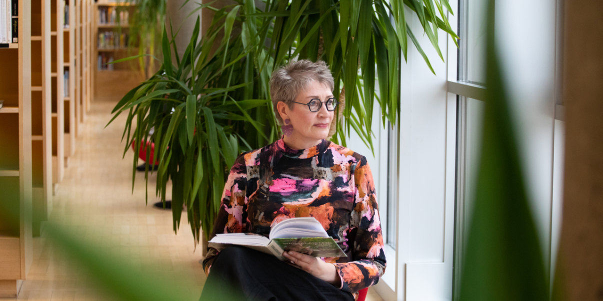 Henkilö istuu kirjastossa kirjahyllyjen ja viherkasvien ympäröimänä, katsoo ulos ikkunasta kirja sylissään.