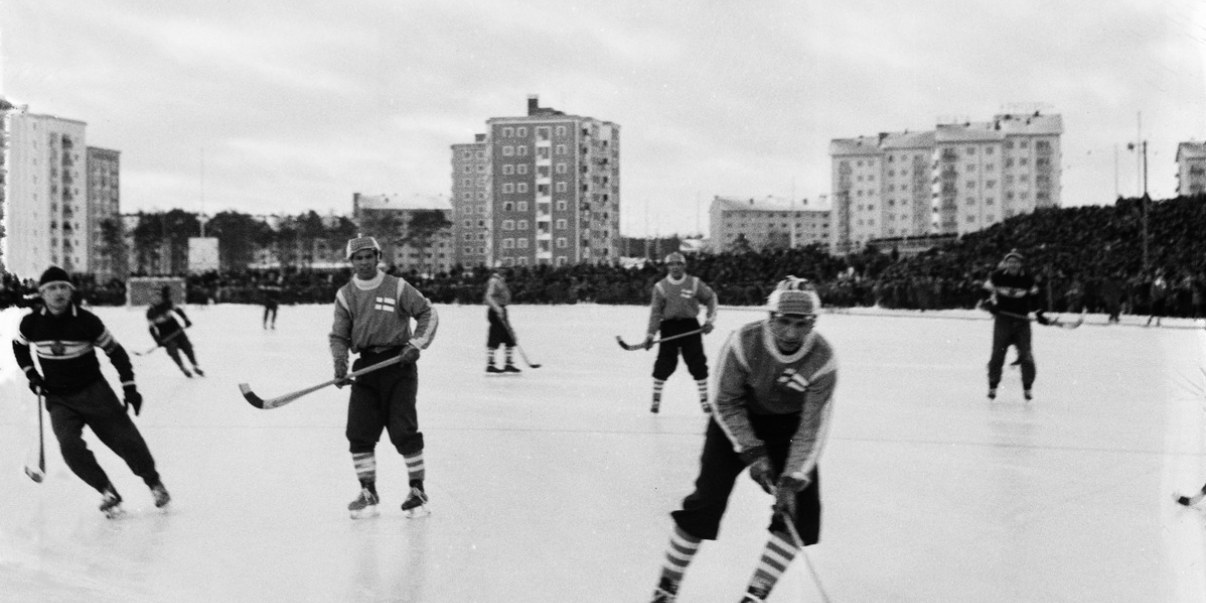 Jääpalloilijoita pelaamassa jäällä, taustalla kerrostaloja.