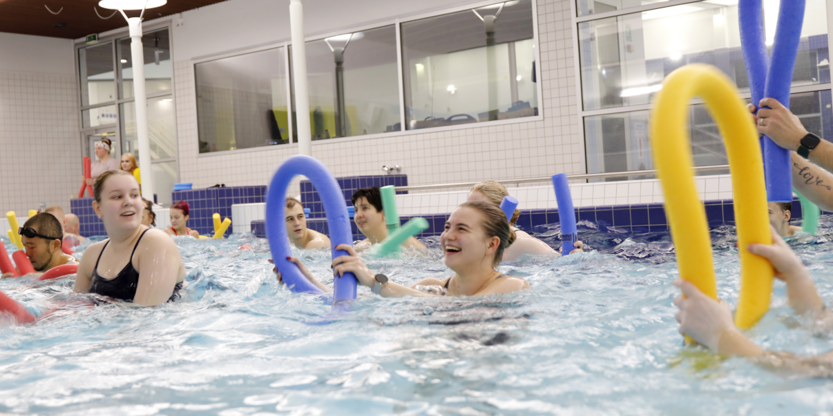 ihmisiä altaassa, iloiset ilmeet, käsissä erivärisiä vaahtomuovipötkylöitä