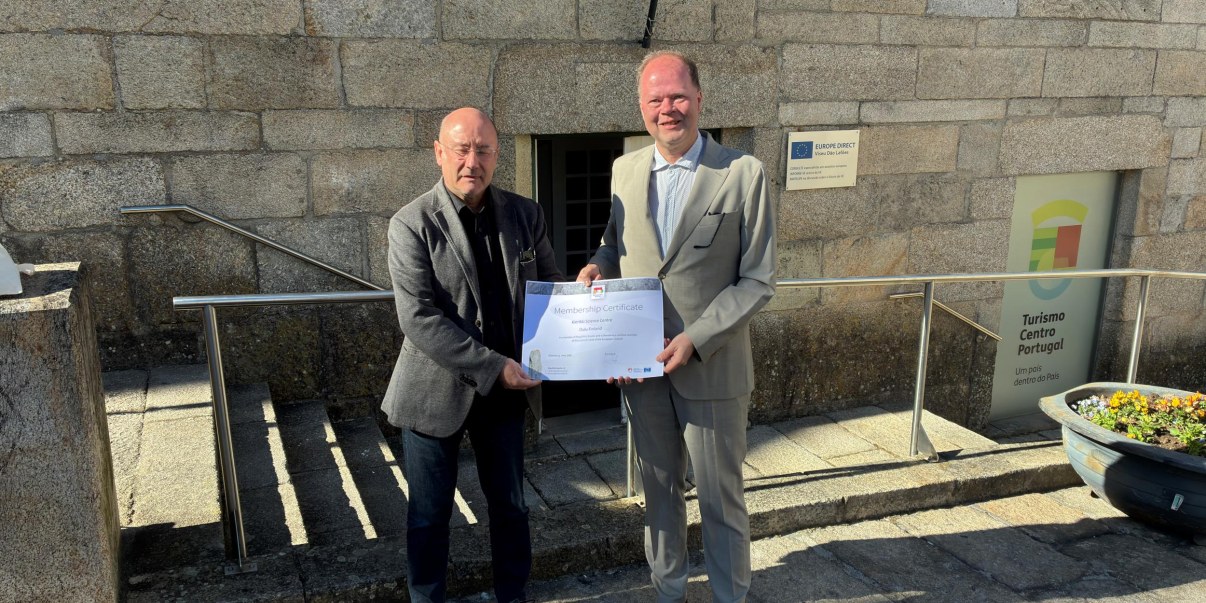 Kaksi miestä harmaissa puvuissa pitelevät yhdessä paperista todistusta.