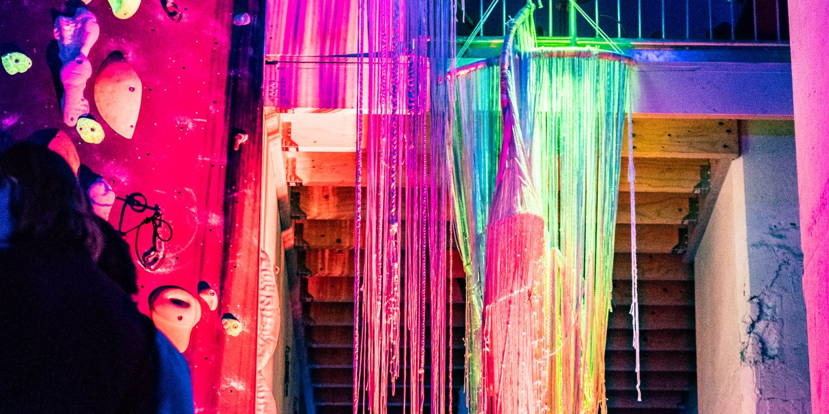 Sateenkaaren väreihin valaistu katosta roikkuva naruteos, taustalla kiipeilyseinä.