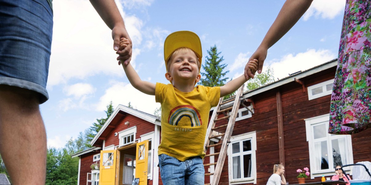 Iloinen lapsi kahden aikuisen välissä, taustalla punainen perinnerakennus.