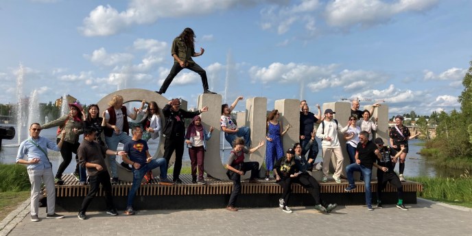 Betonisten OULU -kirjainten läheisyydessä, välissä ja päällä ihmisiä. Soittavat ilmakitaraa, riehakas tunnelma aurinkoisella säällä. Ainolan puiston suihkulähteet taustalla. 