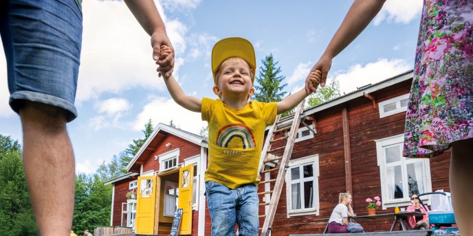 Iloinen lapsi kahden aikuisen välissä, taustalla punainen perinnerakennus.