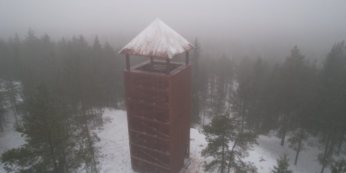 Sumuisessa ja hieman lumisessa metsämaisemassa tumman punainen, korkea puinen näköalatorni. Tornissa katto. Kuva on ilmakuva.