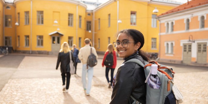 Lyseon piha ja opiskelijoita pihalla, yksi katso kameraan ja muut ovat menossa sisään