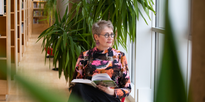 Henkilö istuu kirjastossa kirjahyllyjen ja viherkasvien ympäröimänä, katsoo ulos ikkunasta kirja sylissään.