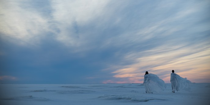 Kaksi henkilöä kävelee jäällä kohti auringonlaskua, heidän päällään oleva kevyt kangas liehuu tuulessa.
