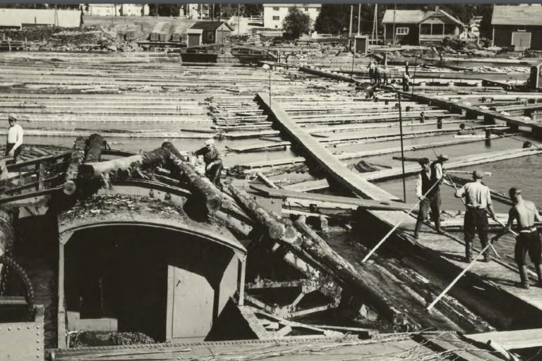 Mustavalkokuvassa uittotyömaa ja ihmisiä sekä rannalla rakennuksia.