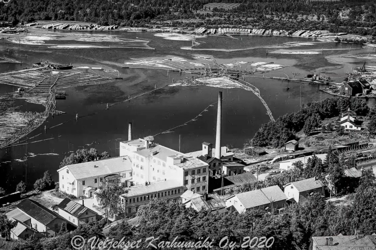 Mustavalkokuvassa tukin erottelua ja rakennuksia jokimaisemassa.
