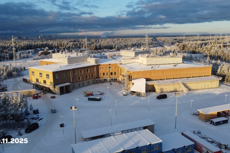Kuvassa lumisessa maisemassa melko valmiin näköinen monitoimitalo auringonvalossa.