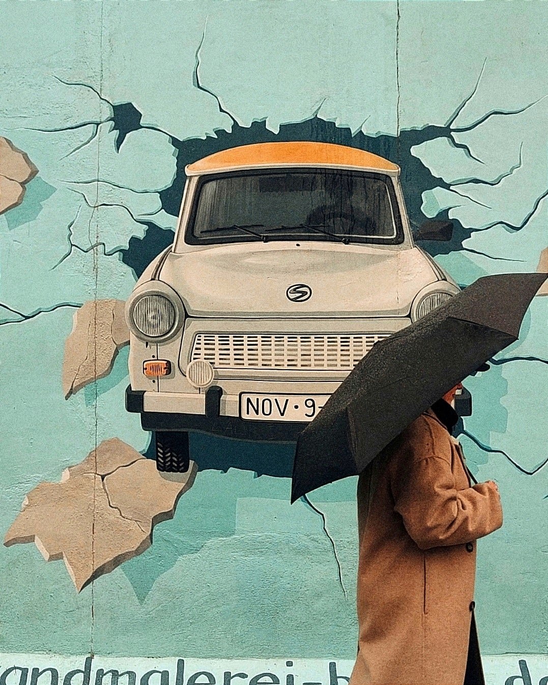 Kuva Berliinin muurilta. Muurin taideteoksessa on valkoinen Trabant-auto. Muurin edessä seisoo henkilö sateenvarjon alla.