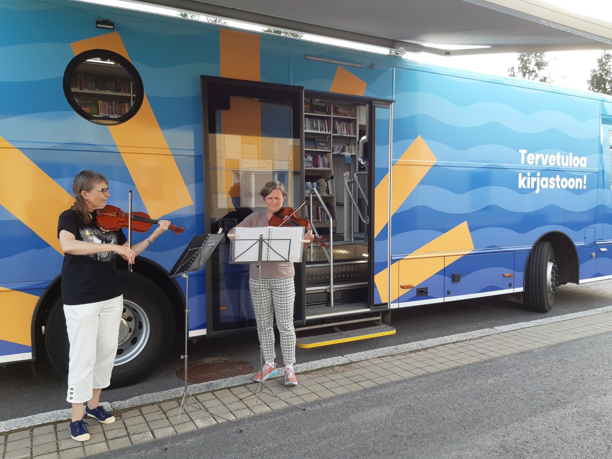 Kaksi henkilöä soittaa viulua kirjastoauton edessä.