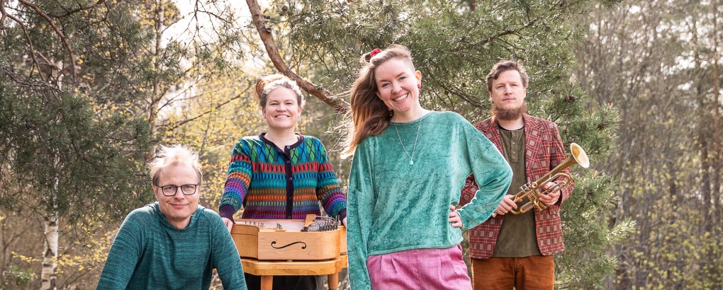 Neljä iloista henkilöä rivissä, yhdellä edessään kannel, toisella kädessään torvi.