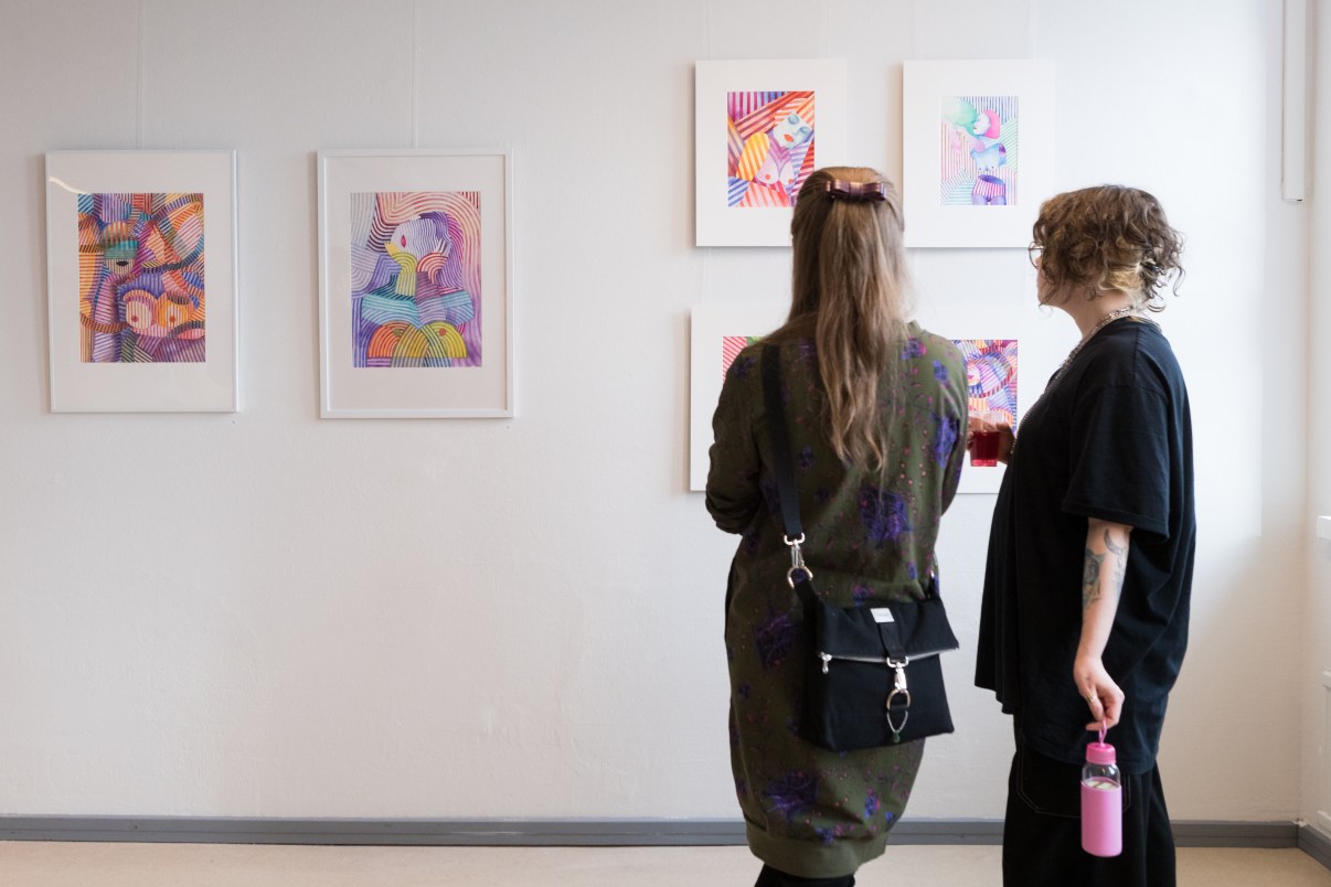 Kaksi henkilöä selät kameraan päin katsomassa valkoisella seinällä olevia värikkäitä akvarellitöitä.