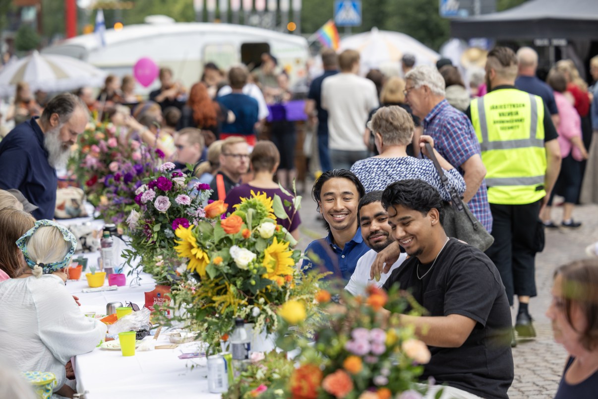 Hymyileviä ihmisiä istumassa pitkän pöydän ääressä ulkona, pöydällä kukka-asetelmia