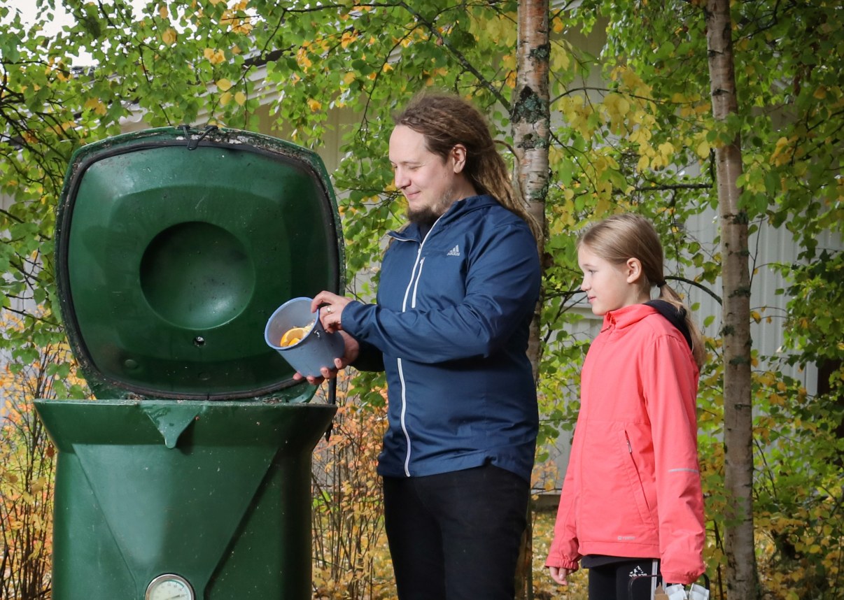 Kuvassa mies ja tyttö seisovat kompostorin vieressä. Kompostorin kansi on auki. Mies kaataa kompostoriin pienestä sankosta biojätettä. Tyttö seuraa sivusta, katsoo kompostorille. 