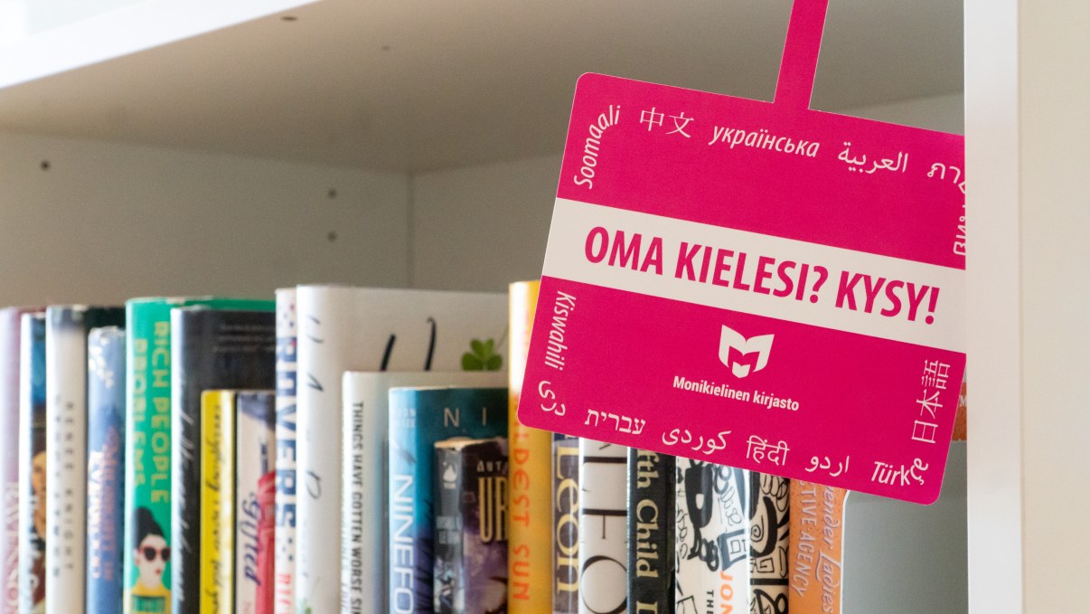 Monikielisen kirjaston mainoskyltti kirjahyllyssä.