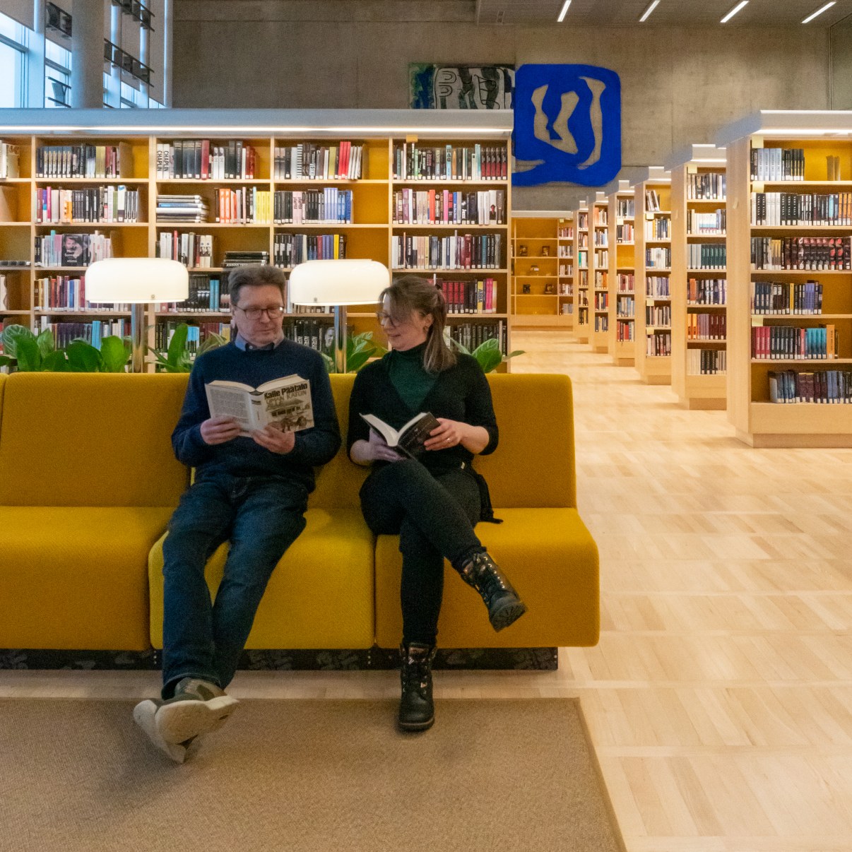 Kaksi henkilöä istuu sohvalla kirjat kädessään, taustalla kirjahyllyjä.