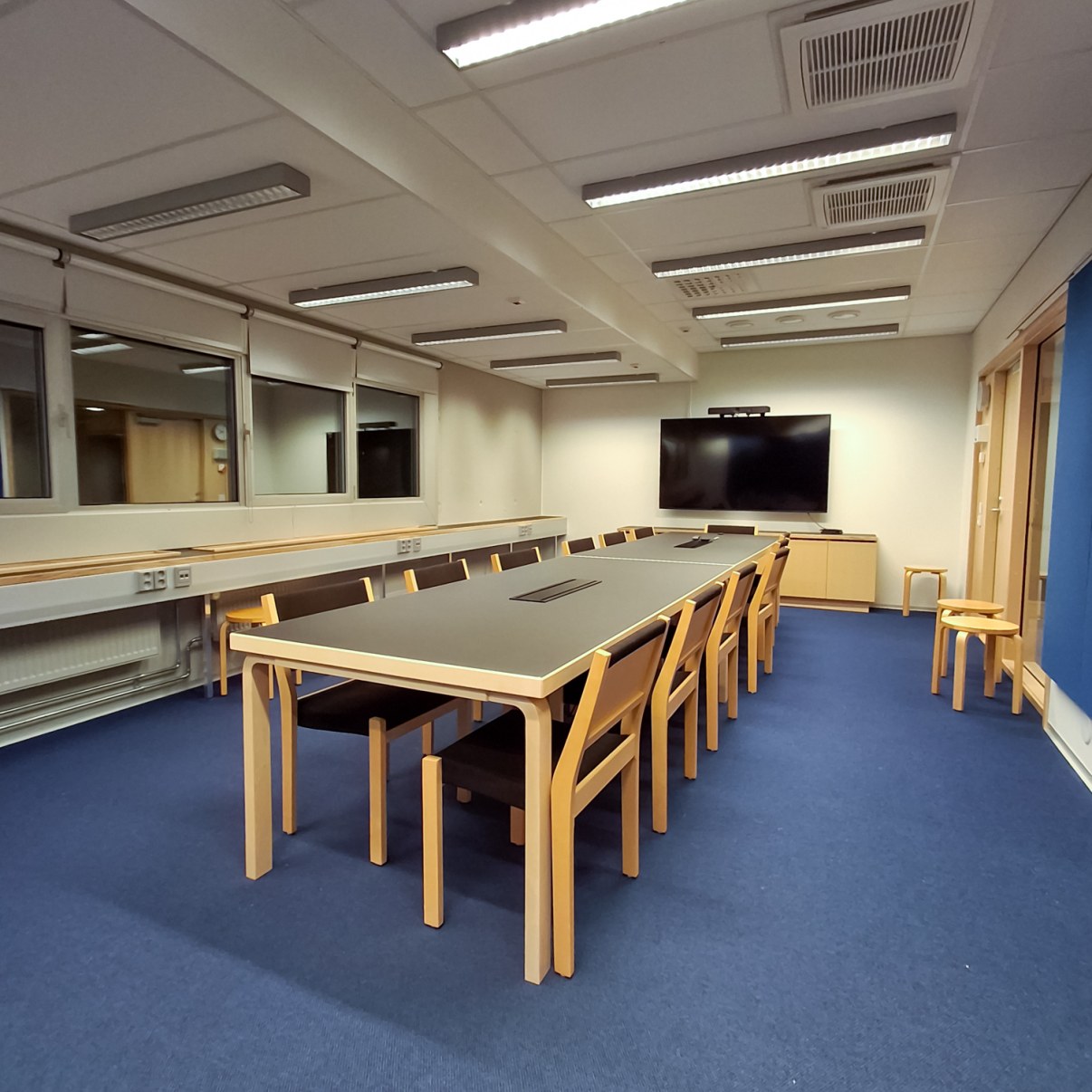 Kokoushuone jossa pöydän ympärillä 12 tuolia, seinällä näyttö.