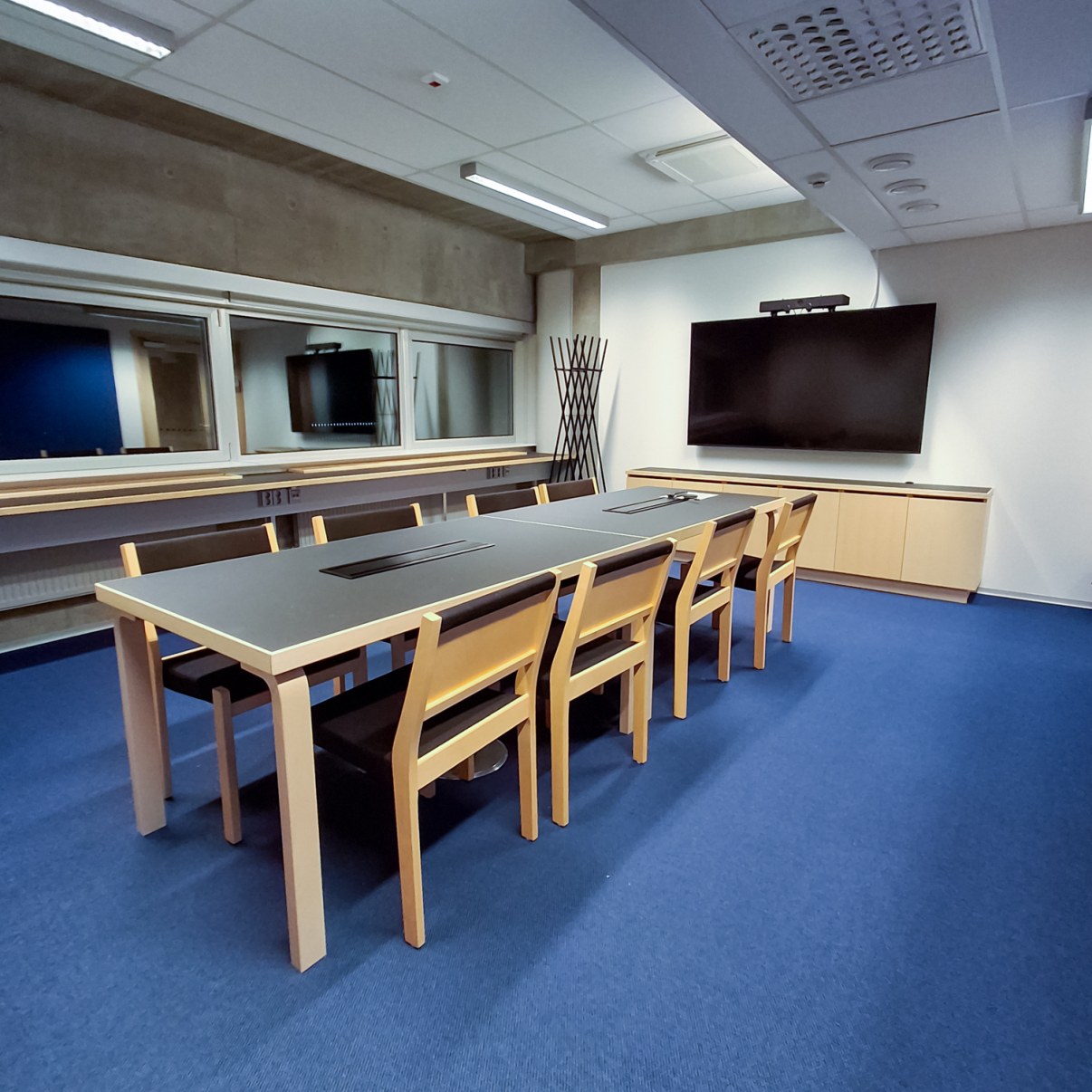 Kokoushuone jossa pöydän ympärillä kahdeksan tuolia, seinällä näyttö.