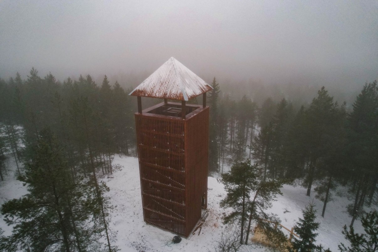 Korkea puinen, tummanpunainen näköalatorni. Maassa vähän lunta, metsää taustalla. Sumuinen keli.