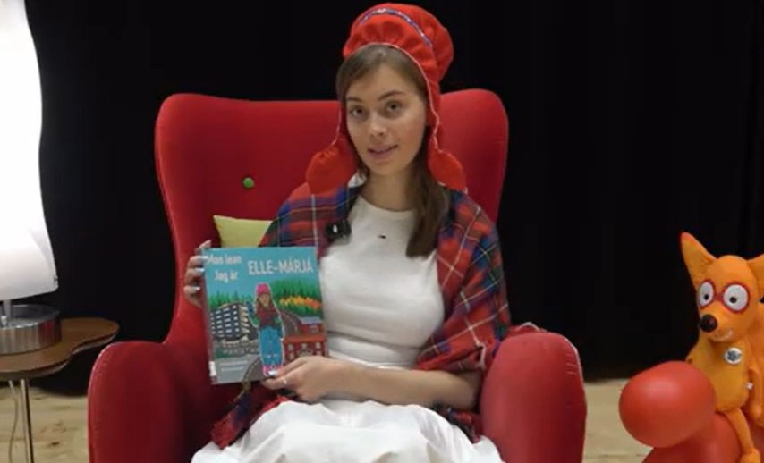 Nainen istuu nojatuolissa ja esittelee luettavaa kirjaa.