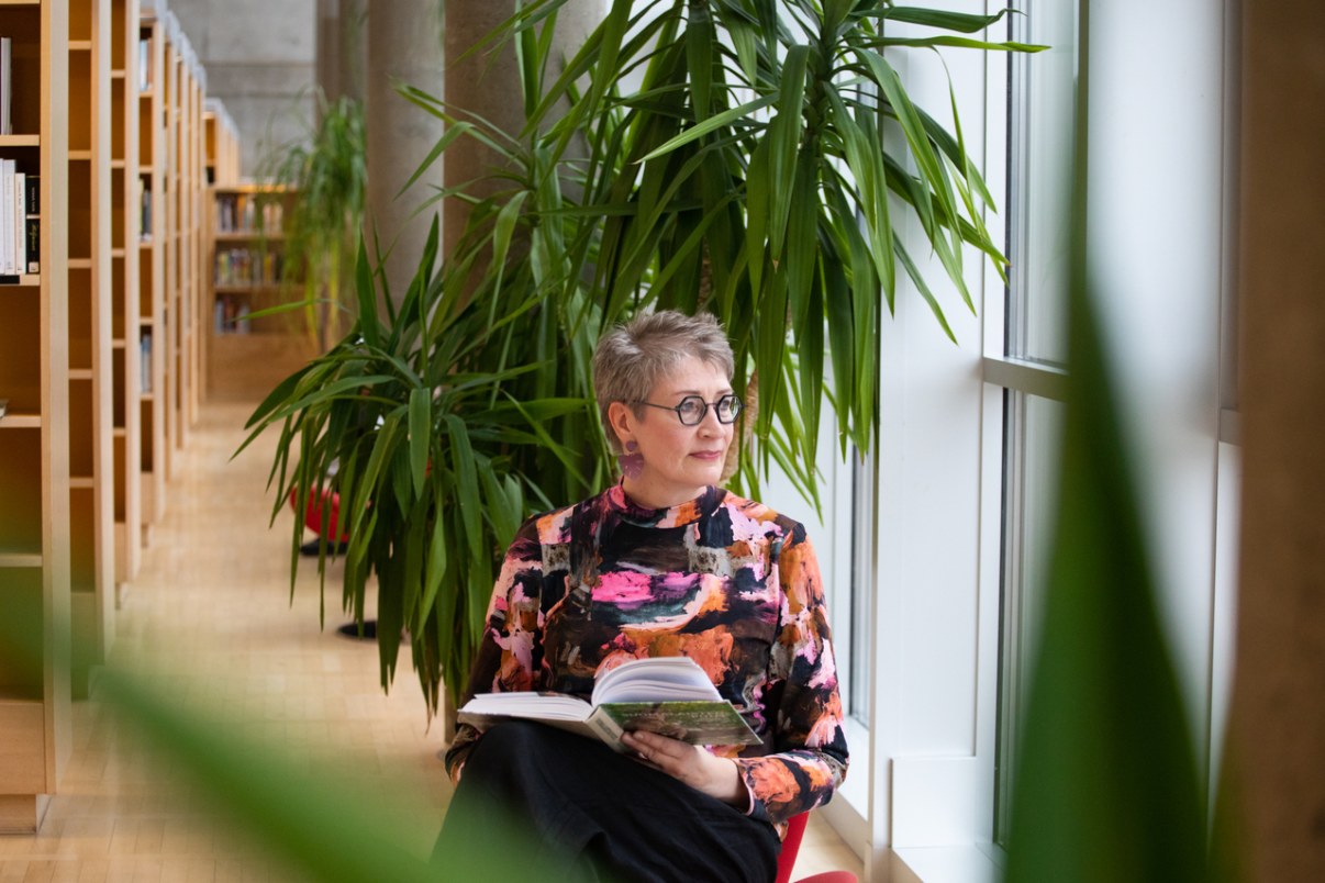Nainen lukee kirjaa kirjastossa ja katselee ulos ikkunasta.