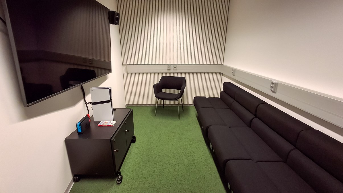 Huone, jossa sohva, nojatuoli, iso televisio seinällä ja sen alla pieni hyllykkö.