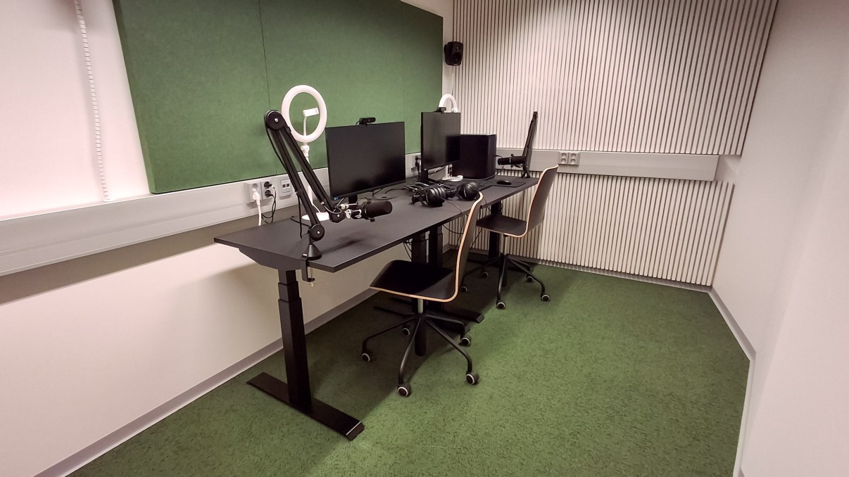 Huone, jossa pöytä jolla kaksi näyttöä, tietokone ja erilaisia kuvaus- ja äänitystarvikkeita.