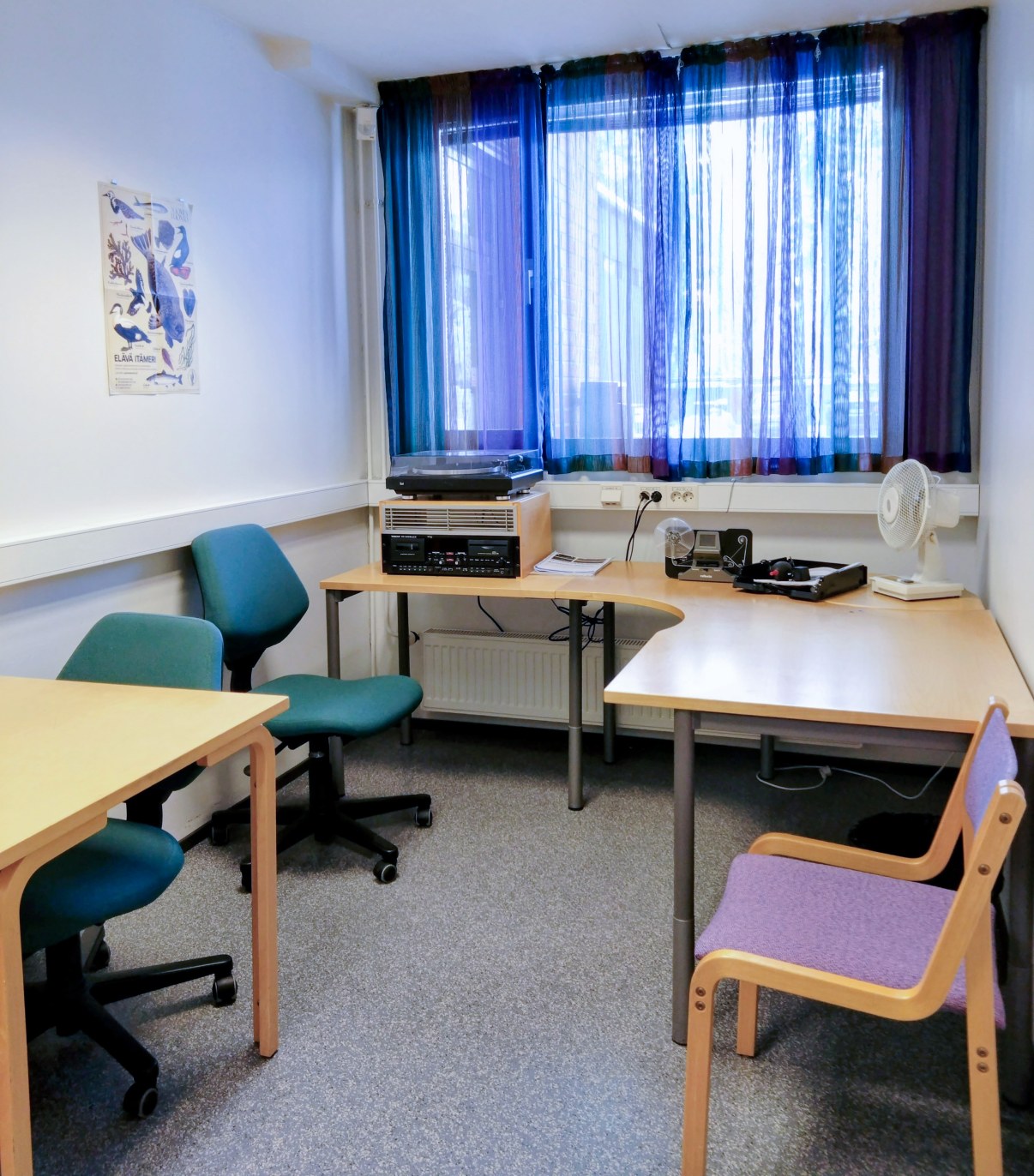 Näkymä työtilaan jossa kaksi työpöytää, kolme tuolia ja erilaisia laitteita.