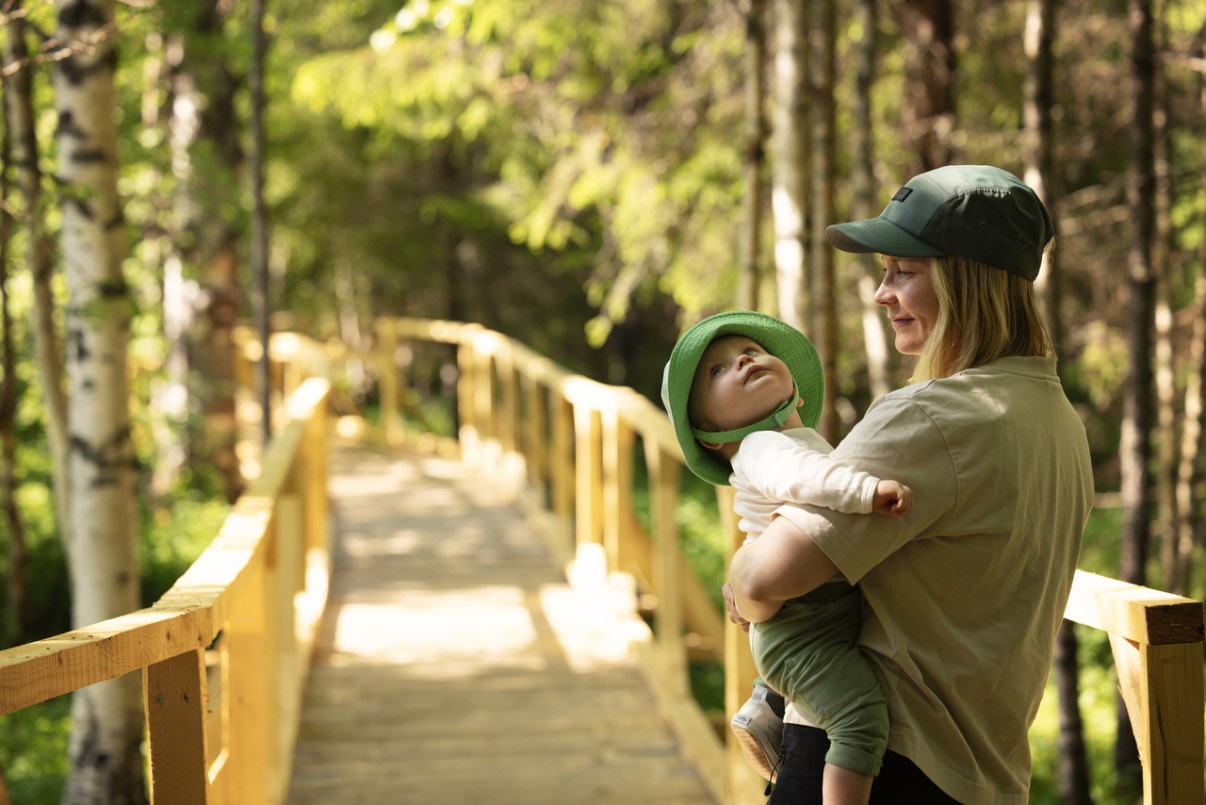 Äiti pitää pientä lasta sylissään. Esteetön, puinen reitti jatkuu taustalle metsään.
