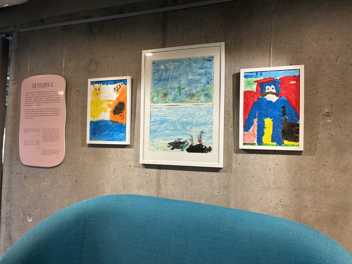 Kolme lasten tekemää kehystettyä taideteosta betoniseinää vasten. Teosten vieressä tekstikyltti, jossa on lasten kirjoittamia tarinoita. 