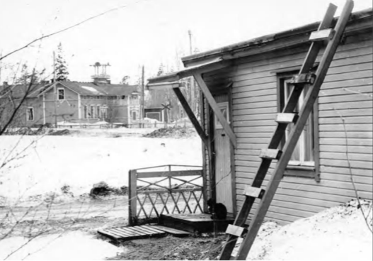 Mustavalkoisessa kuvassa pieni puutalo terasseineen ja tikapuineen. Taustalla siintää huvilarakennus.