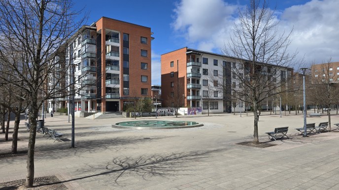 Kuvassa kerrostaloja, puistonpenkkejä ja suihkulähde.