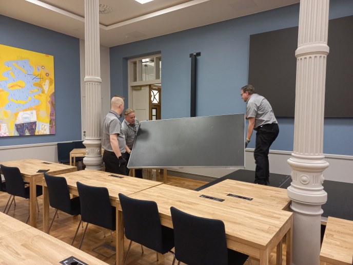 kaksi virastomestaria nostaa pöytää kaupungintalon valtuustosalissa. Yksi seuraa vieressä.