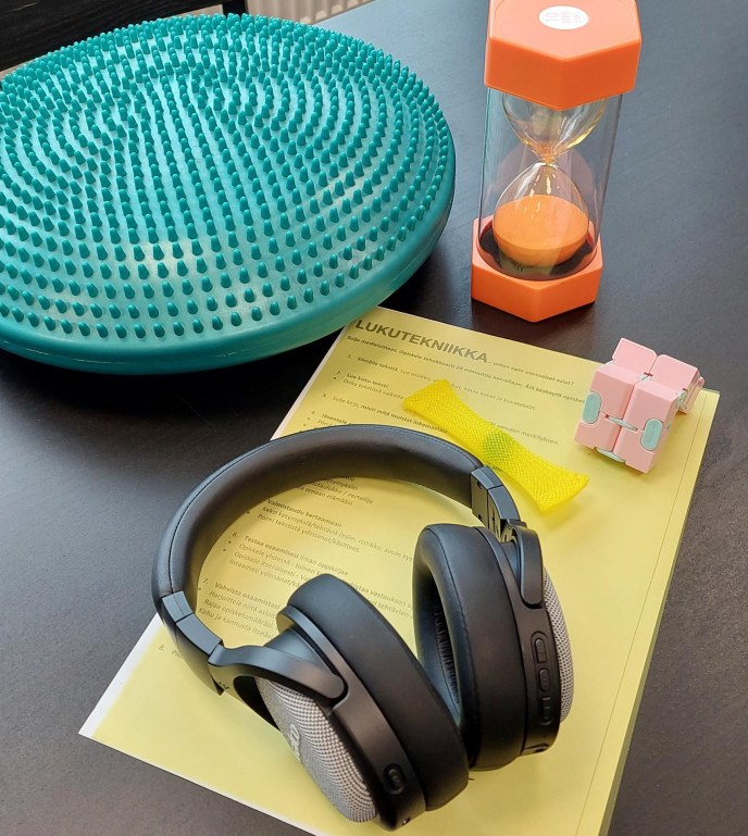 Pöydällä kuulokkeet, tasapainolauta, tiimalasi ja erilaisia stressileluja.