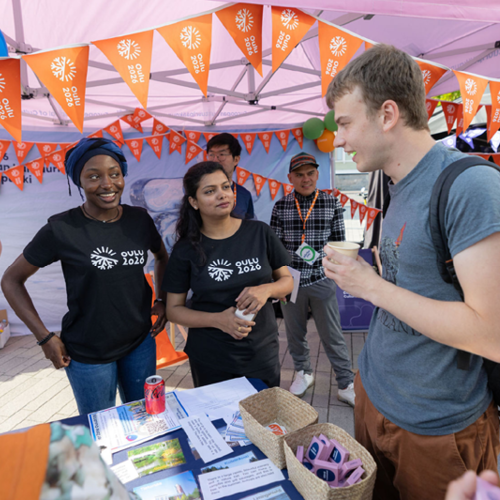 Kaksi ihmistä seisoo pöydän takana ja yksi pöydän edessä, ihmiset keskustelevat. Taustalla ja etualalla näkyy oranssia lippuviiriä ja vaaleanpunainen telttakatos.