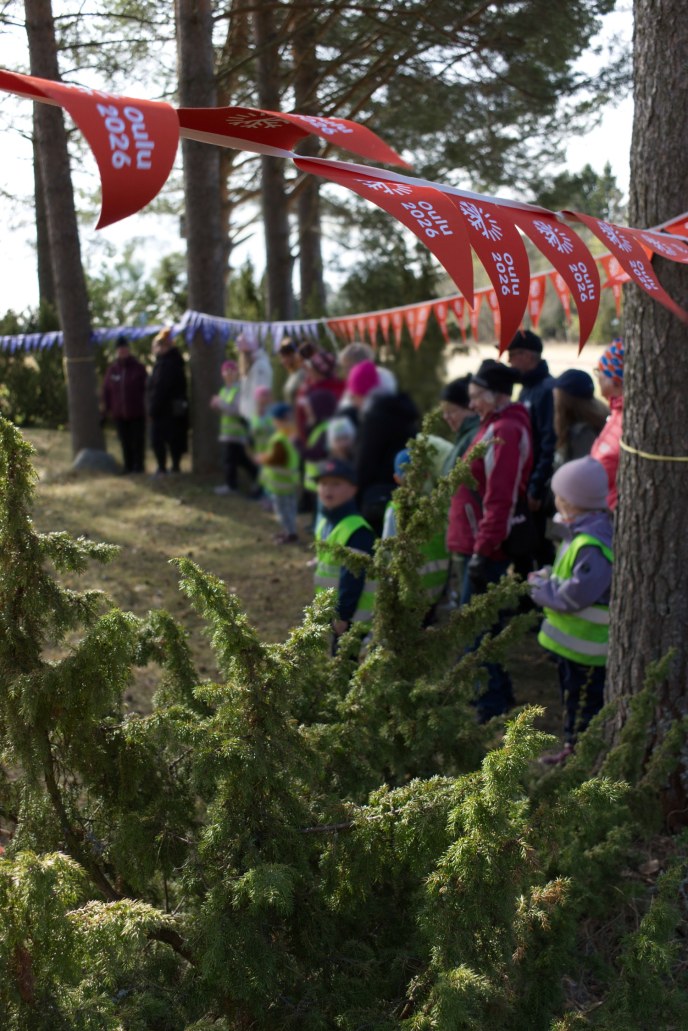 etualalla vihreitä kuusenoksia ja taustalla ihmisiä seisoo rivissä puiden välissä. Heidän yläpuolellaan pitkä viirinauha. Viireissä lukee: Oulu2026.